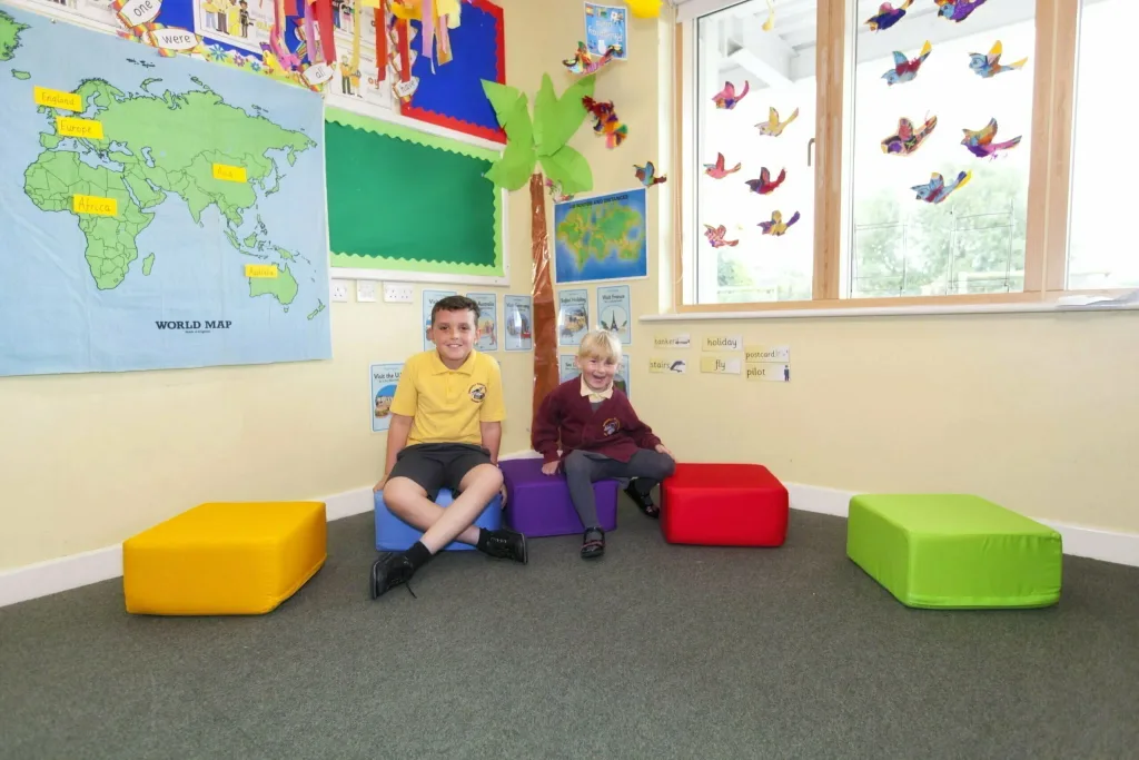 Five cotton foam seating pods (yellow, blue, purple, red, green) with two children on blue and purple in a classroom by a WORLD MAP.
