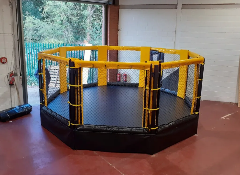 Octagonal steel training cage with black padded base, yellow padded top rails and chain-link mesh sides on a raised deck.