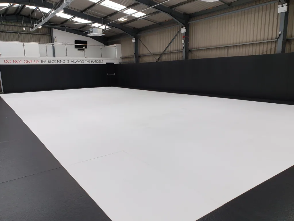 White PVC combat mat with black border in a training hall, with text "DO NOT GIVE UP THE BEGINNING IS ALWAYS THE HARDEST" on the far wall.