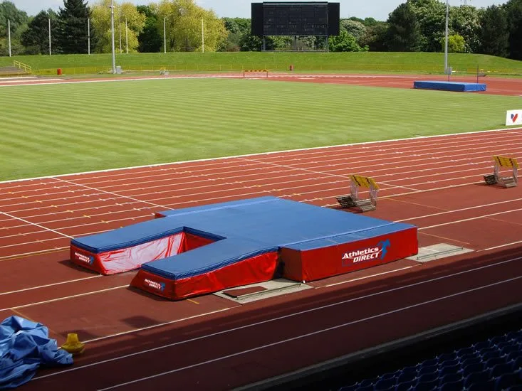 Blue-topped red landing mat with a U-shaped cutout on a red running track beside marked lanes and a green infield with a scoreboard.