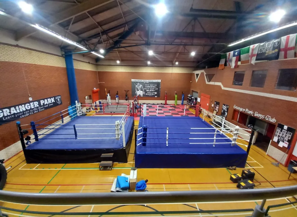 Two adjacent raised boxing rings with blue canvases and white ropes on a gym floor under high rafters, with a wall sign reading "Grainger Park Boys Club".