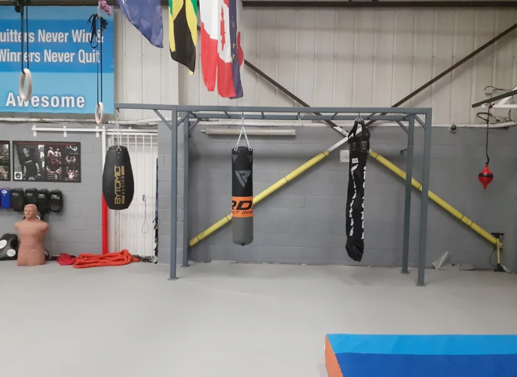 Free-standing steel equipment rig, powder coated, holding three hanging punching bags in a gym with a wall sign reading "Awesome".