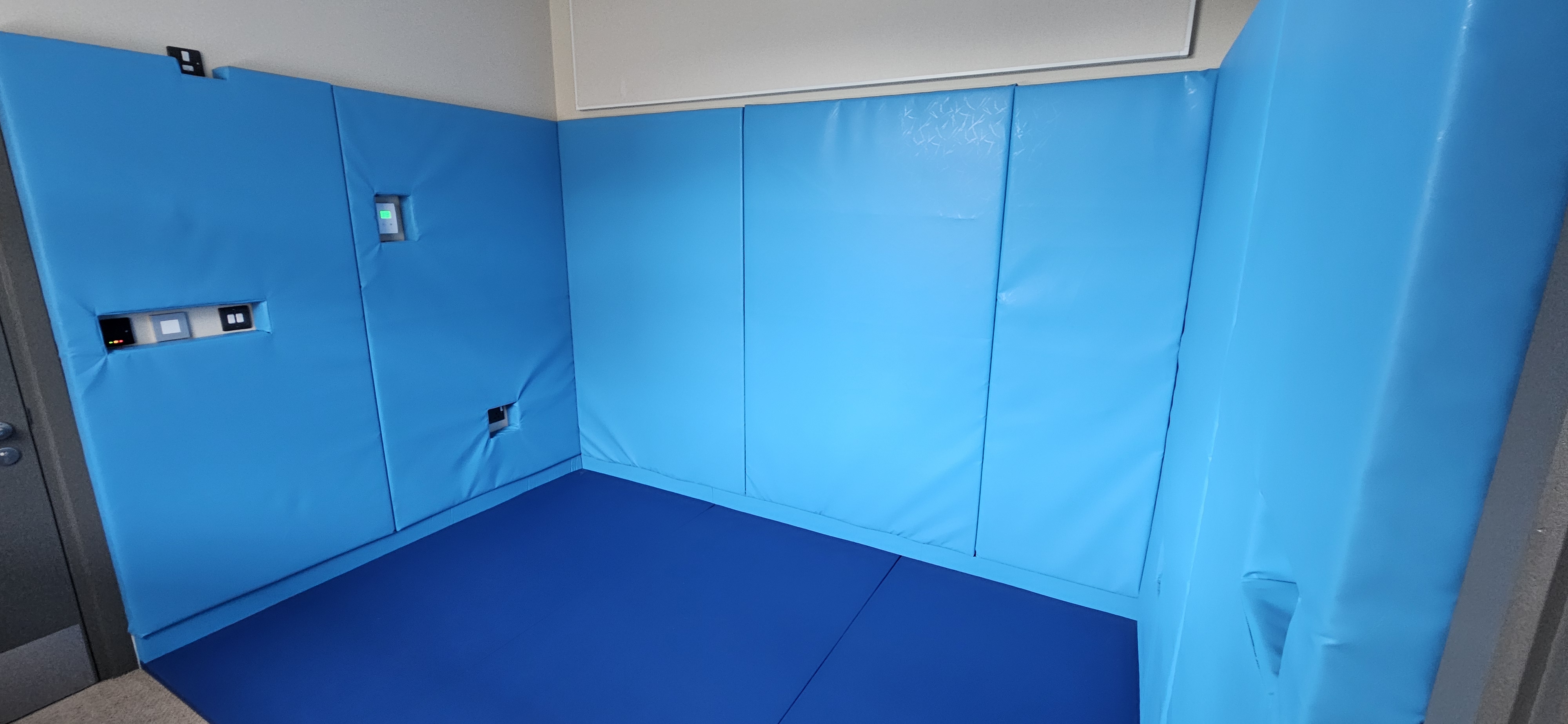Wall padding Corner room with bright blue padded wall panels and matching blue floor mats with cutouts for outlets and controls.
