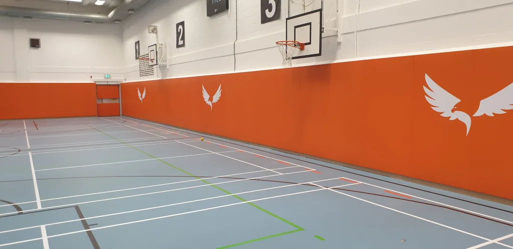 Gym hall with orange wall padding bearing white wing logos, basketball hoops, blue floor court lines, and black panels labeled 2 and 3.