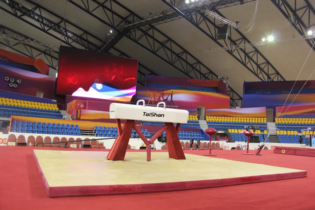 Competition pommel horse with red genuine leather body over wood and foam, steel base and two handles, on a red mat in an empty arena.