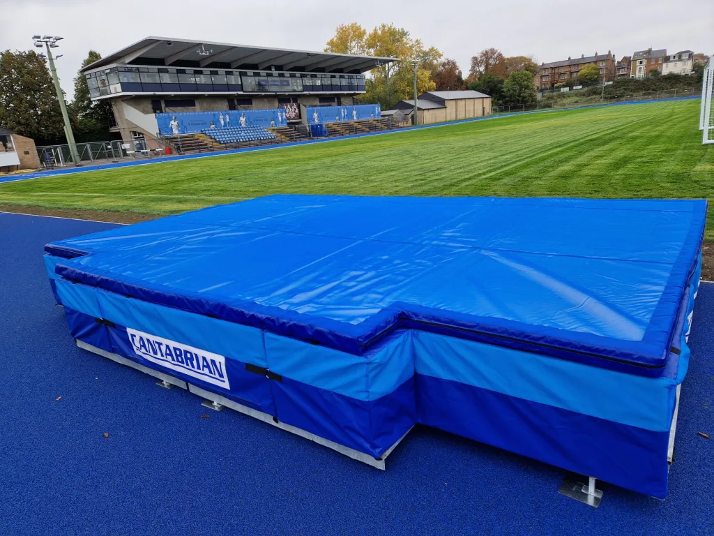 Blue high jump landing mat with CANTABRIAN label and heavy-duty UV stabilised PVC cover on a blue track beside a grass field.
