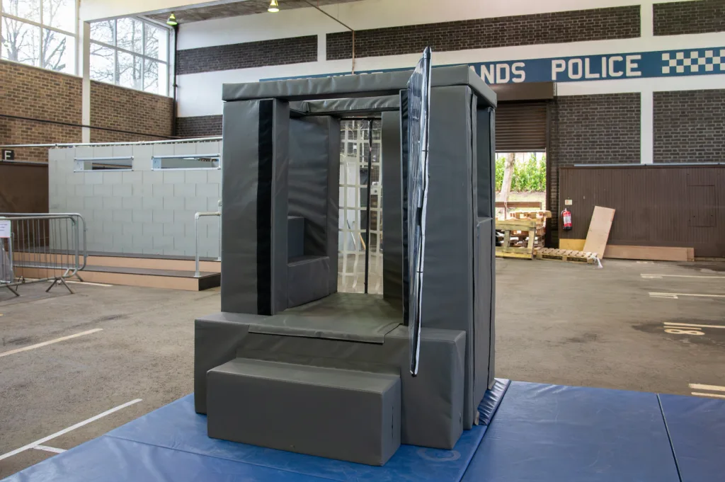 Gray reinforced-foam van cage with a heavy-duty PVC cover and step entrance on a blue mat in a training hall, wall sign reads "ANDS POLICE".