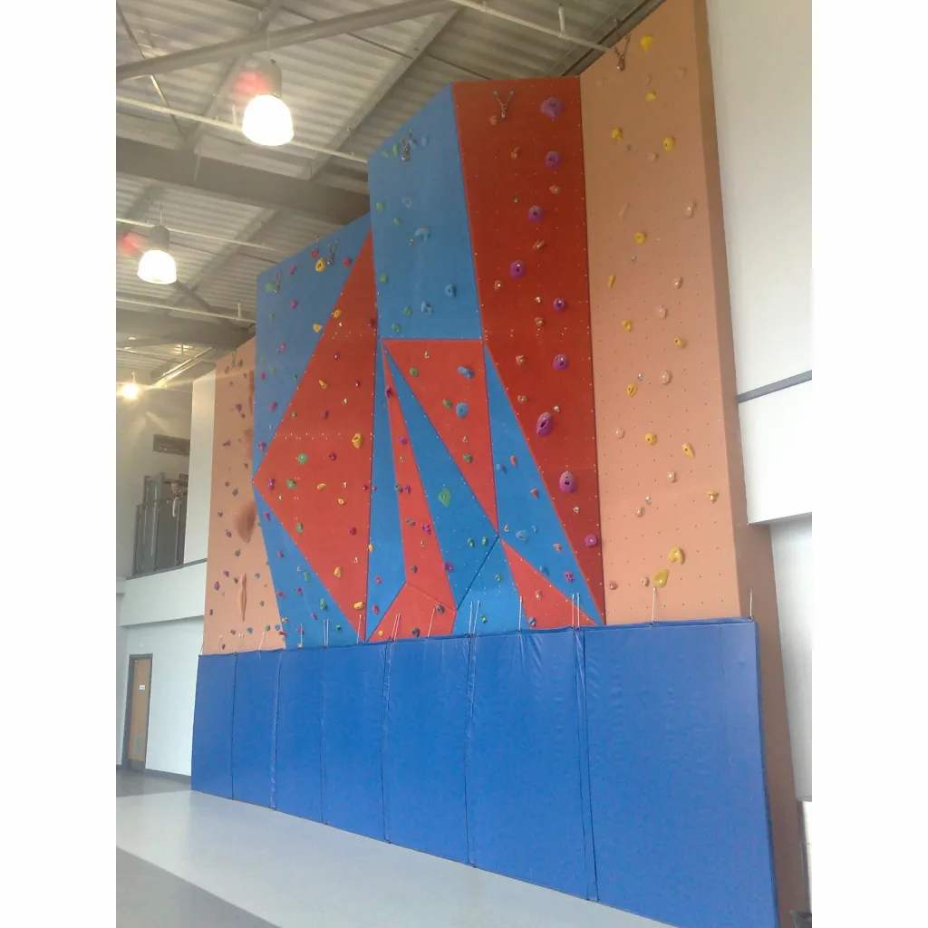 Blue articulated mats folded upright along the base of a tall indoor climbing wall with red and blue panels and multicolored holds.
