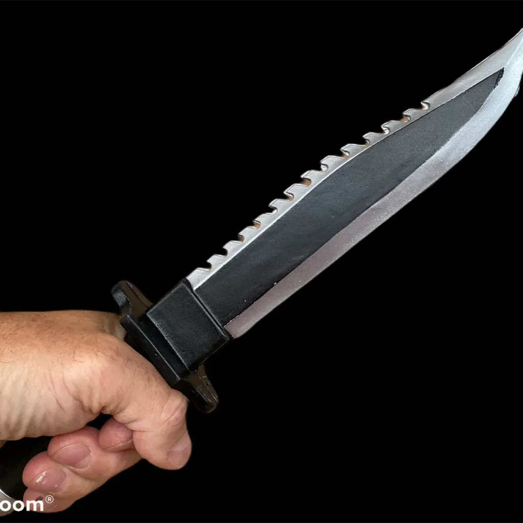 Hand grasping a black and silver serrated survival knife training prop made from PU foam, with small text "oom®" visible.