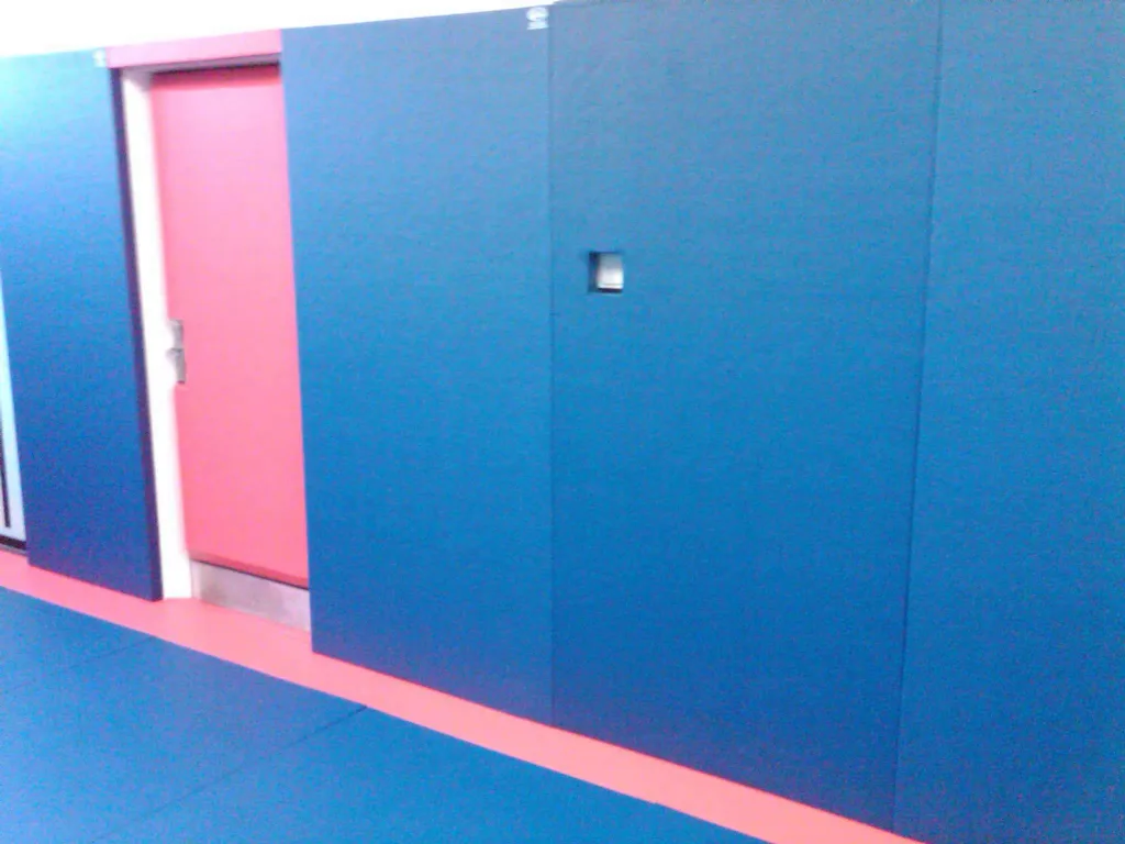 Blue padded wall panels with a small recessed handle beside a red door and red floor border over blue floor mats.