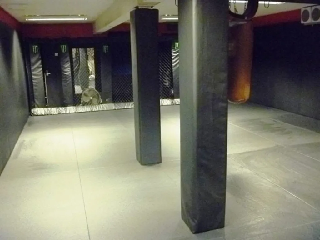Two black polyurethane foam square post pads covered in heavy duty PVC around vertical posts in a padded training room with mats and a hanging punching bag.