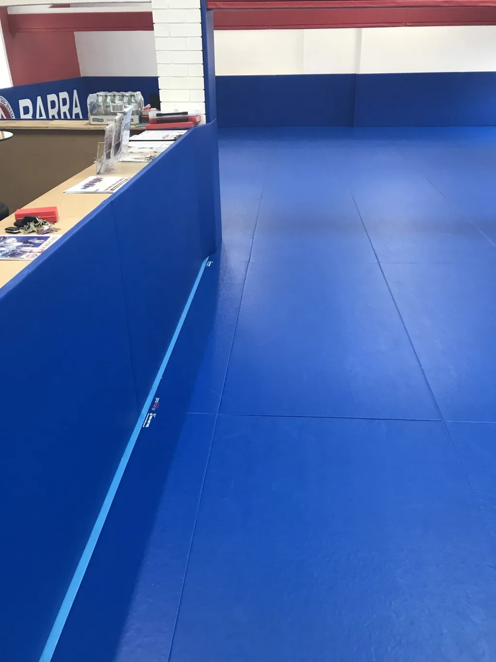 Royal blue BJJ mats covering a training area with padded perimeter and reception counter, sign text 'BARRA' visible.