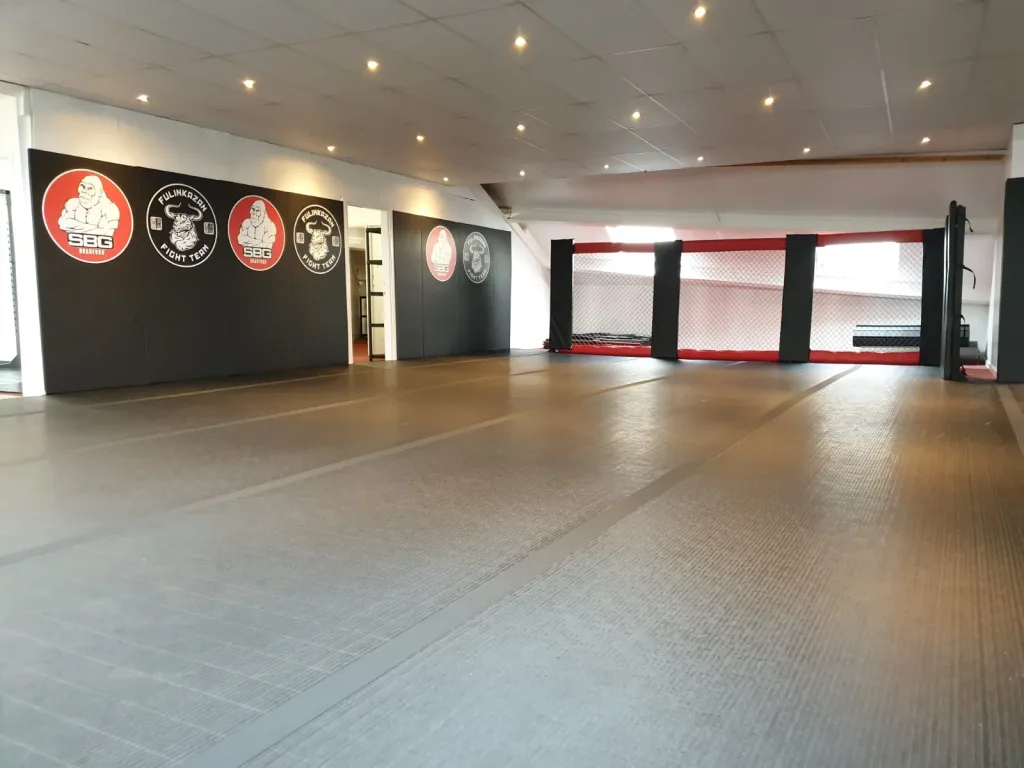 Large grey roll-out mats covering a martial arts training room with padded walls showing 'SBG' logos and a red and black cage at the far end.