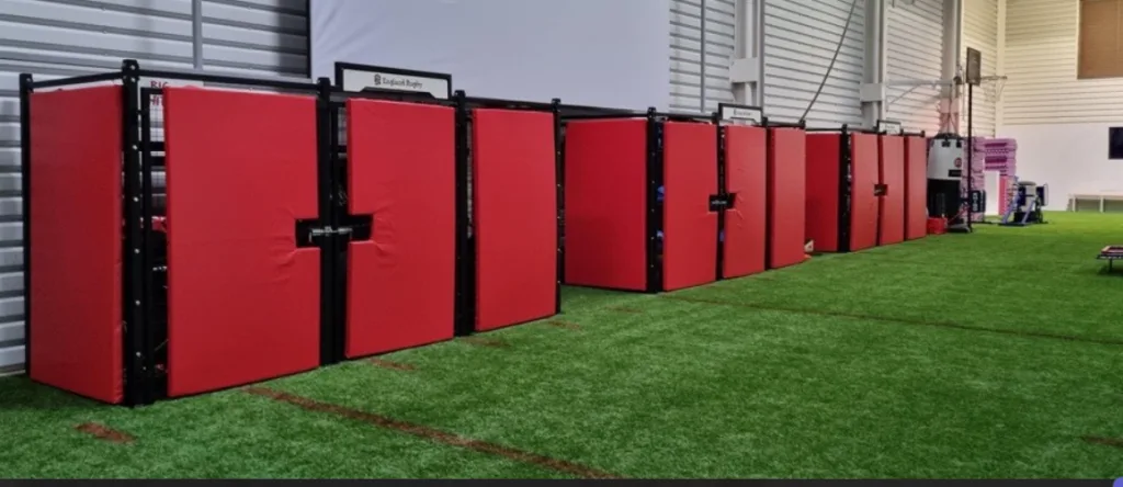 Row of red polyester PVC wall pads mounted on black frames along an indoor sports hall wall above artificial turf.