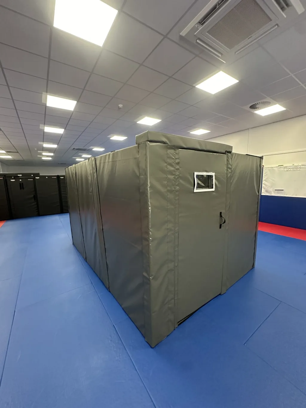 Gray PVC-covered foam custody cell with a small rectangular window and door handle on blue training mats in a spacious practice room.