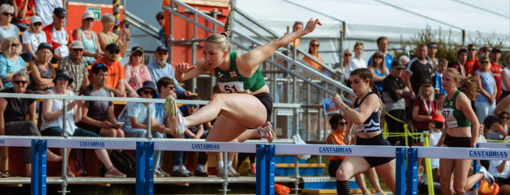 Female hurdler in a green singlet leaping over a hurdle labeled CANTABRIAN, with competitors and a seated crowd behind her.