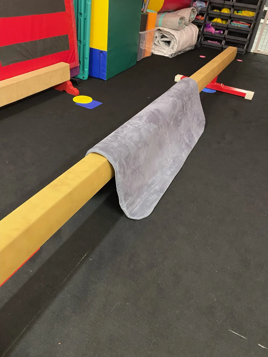 Grey soft jersey balance beam mat with shock-absorbing PU foam core and anti-slip base draped over a balance beam on a black gym floor.