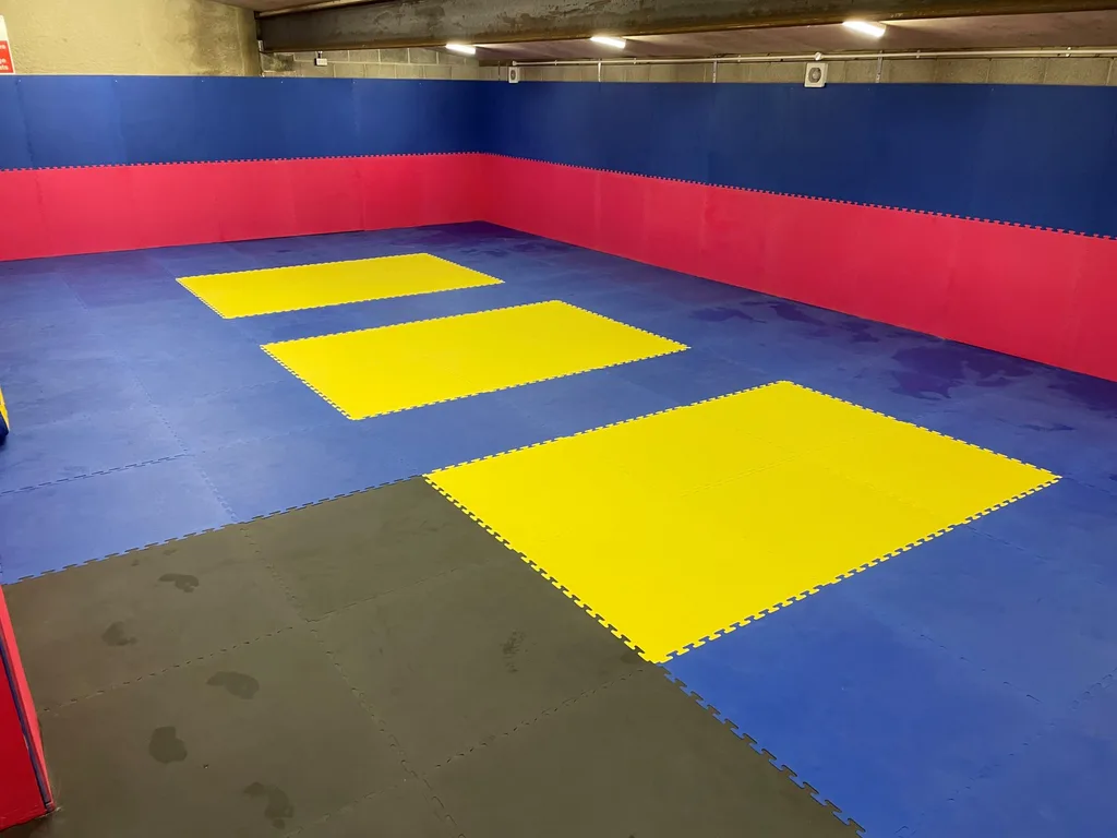 Training room with blue interlocking floor mats, a red padded wall band, three yellow square practice mats centered, and a black mat area in the foreground.