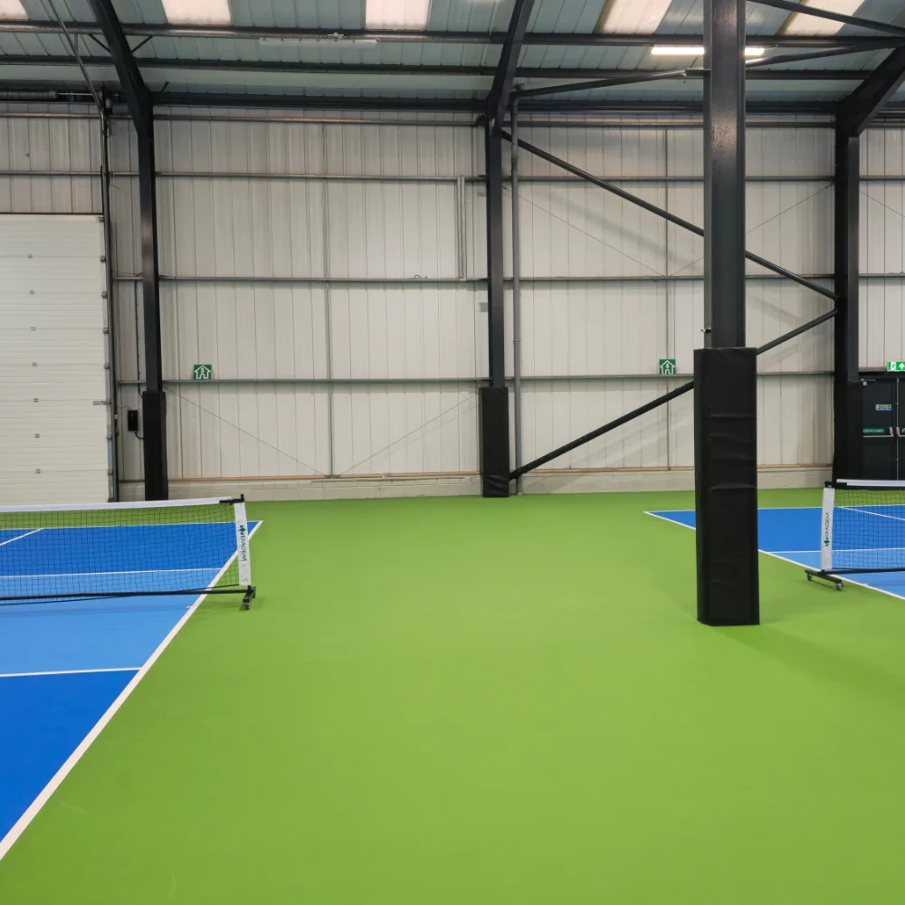 Black PVC-covered high-density foam padding on steel posts beside blue pickleball courts with a green surrounding floor inside an indoor sports hall.