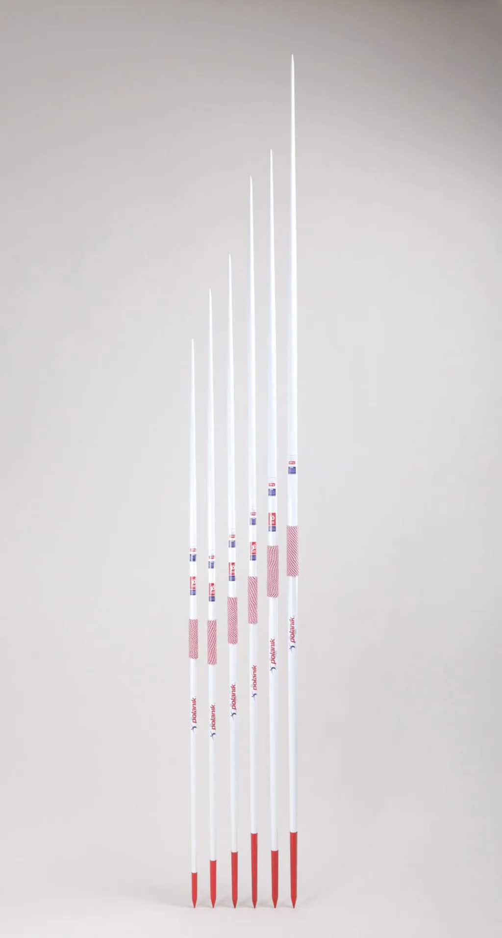 Six white aluminium school javelins of increasing length with red hand grips and red steel tips standing upright.