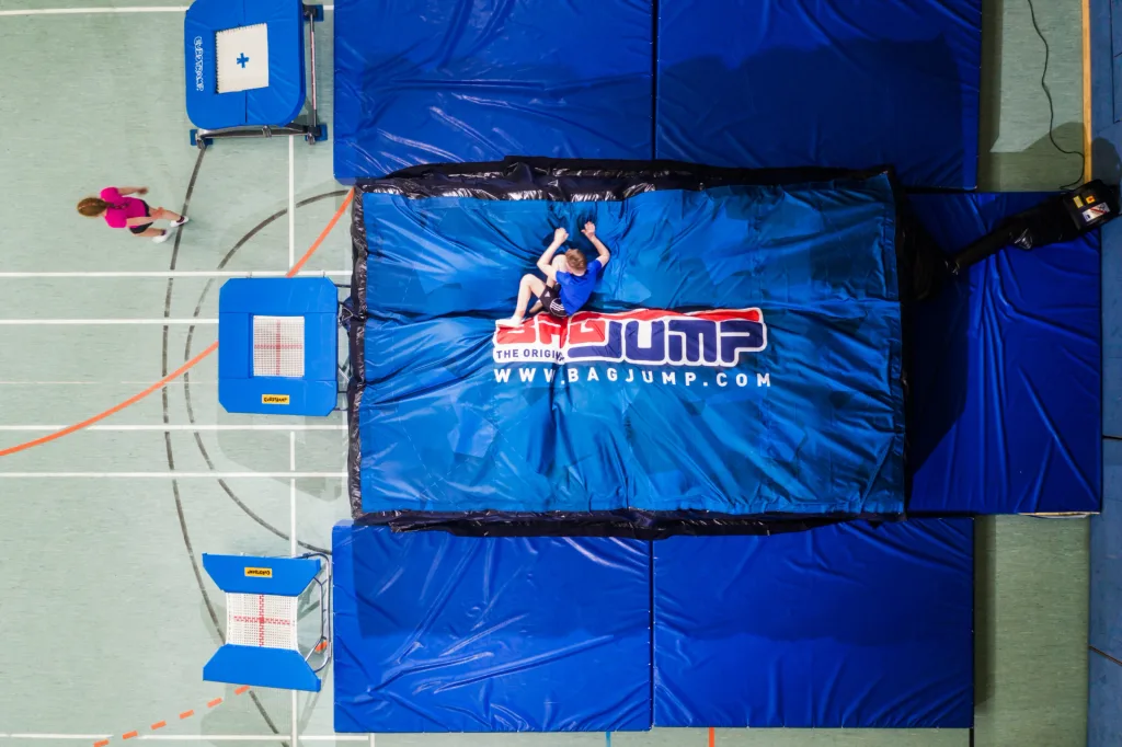 Boy in blue shirt lies on a large blue inflatable mat labeled 'THE ORIGINAL WWW.BAGJUMP.COM' inside a gym.