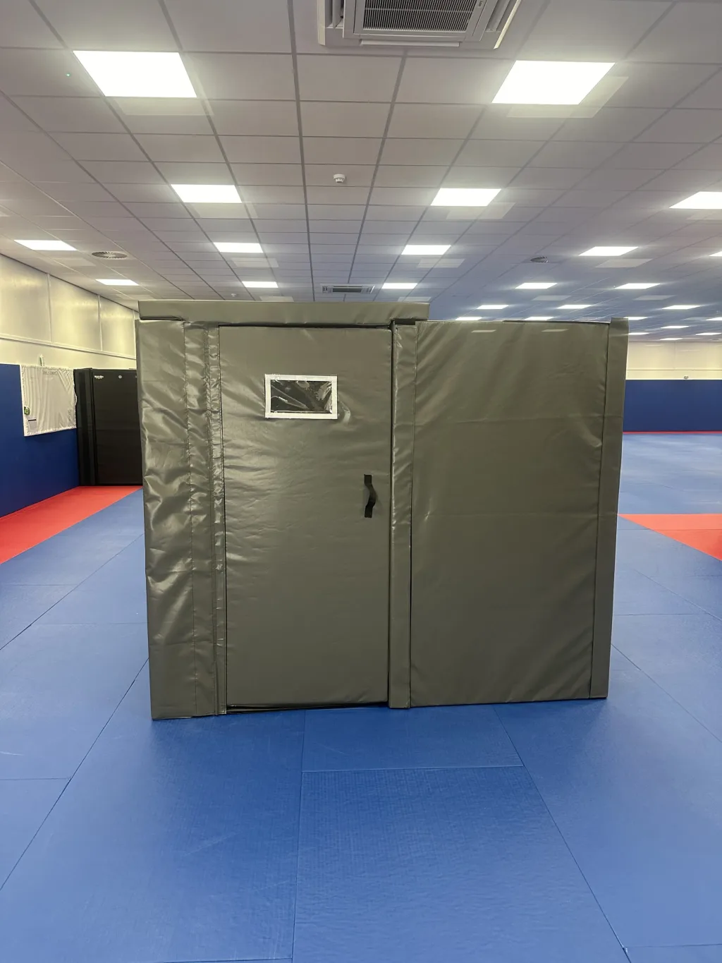Foam custody cell wrapped in heavy-duty PVC with a small observation window and strap handle, set on training mats inside a large hall.