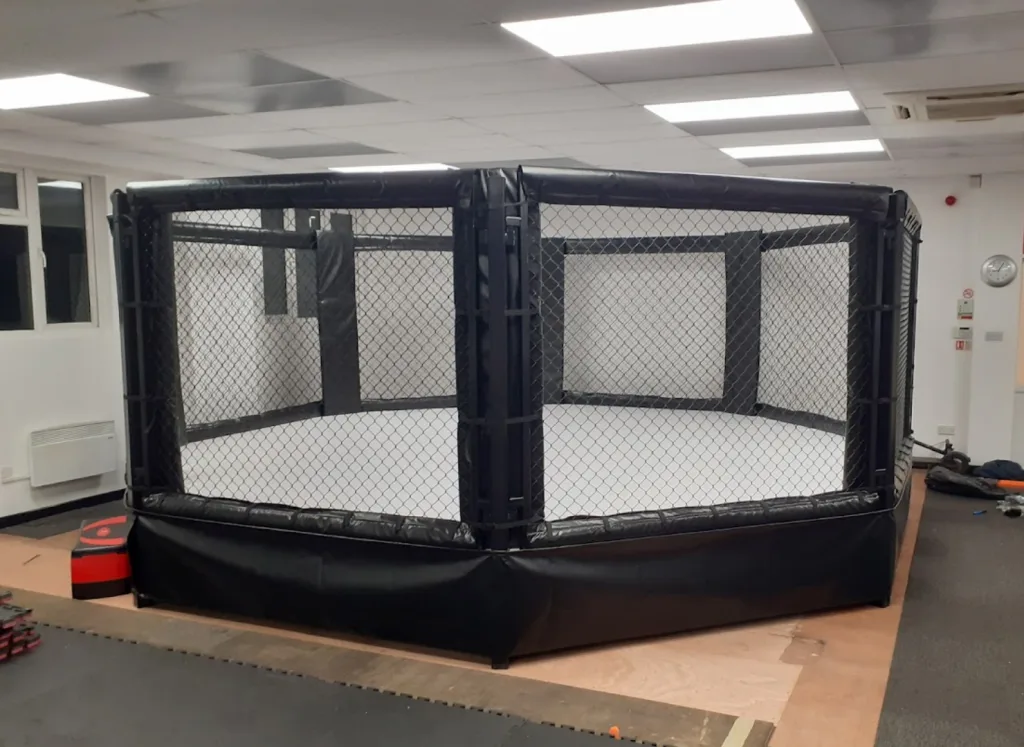 Raised powder-coated steel training cage with chain-link mesh, padded top rail and skirt and a white interior deck inside a gym room.