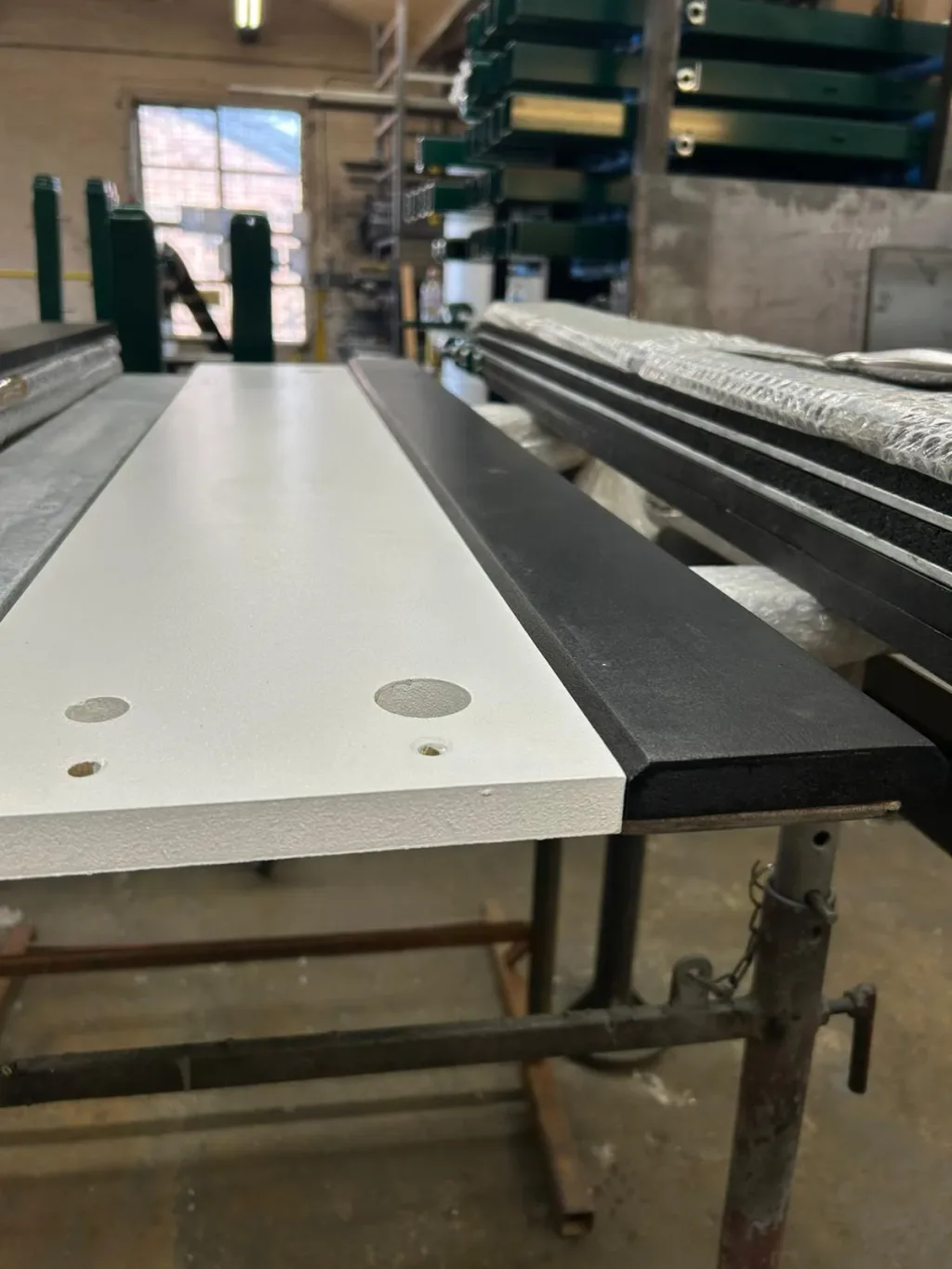 White wooden take-off board with pre-drilled holes and a black rubber edge resting on galvanized steel supports in a workshop.
