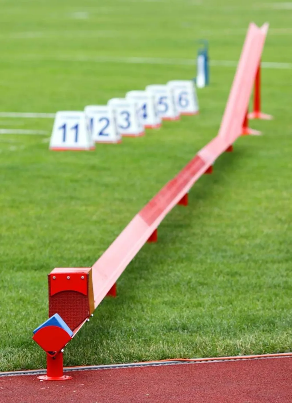 Red shot put return channel made of galvanised steel segments on a grass field with numbered markers "11 12 3 4 5 6" in the background.