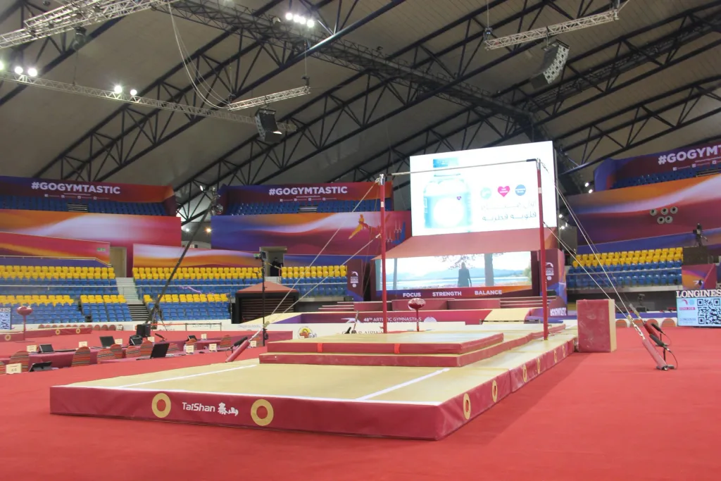 Competition uneven bars with beech-veneer fiberglass rails mounted above red layered landing mats in an empty indoor arena with yellow and blue seating.