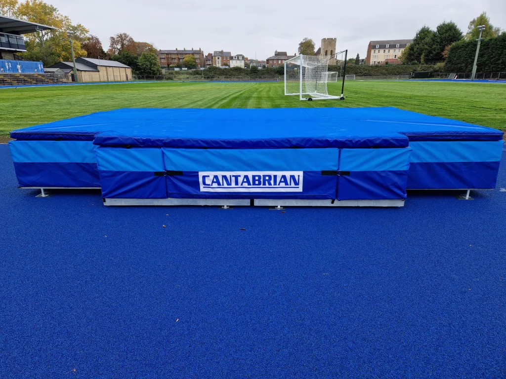 Blue landing mats on a galvanised steel high jump platform labeled CANTABRIAN, on a blue track with grass field and goal behind.