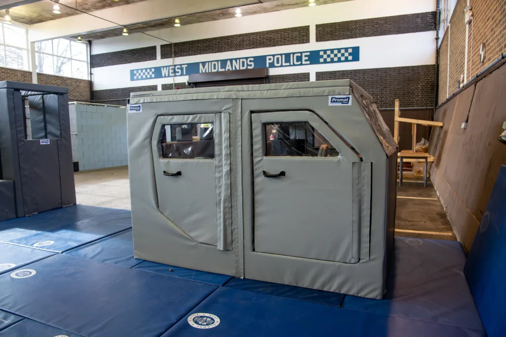 Grey PVC-covered training car with windowed doors and handles on blue mats beneath a sign reading WEST MIDLANDS POLICE.