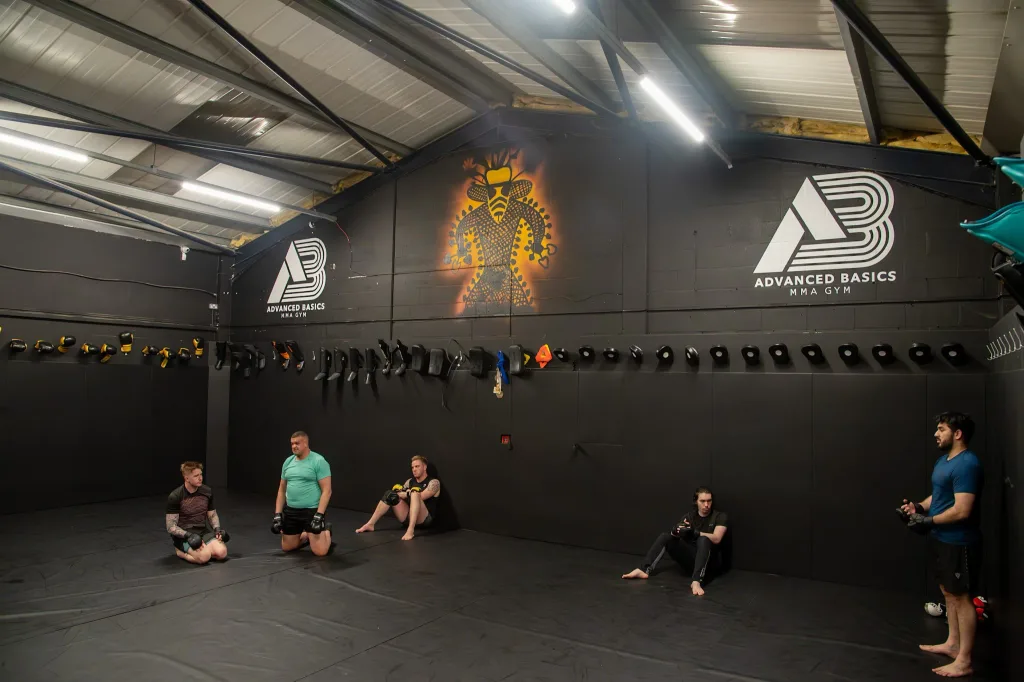 Black vinyl mat cover on gym floor and walls in an MMA training room with people resting, wall logo reading ADVANCED BASICS MMA GYM.