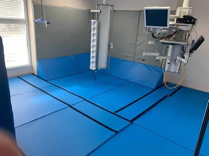 Blue padded floor mats covering the room meet gray padded walls with a mounted medical monitor and ceiling equipment near a window.