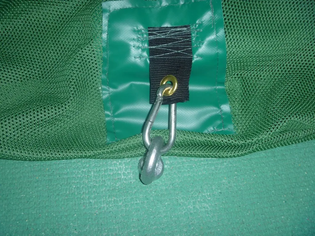 Close-up of a green mesh cage panel with a vinyl patch and black strap with brass grommet holding a silver carabiner and metal anchor on a green mat.