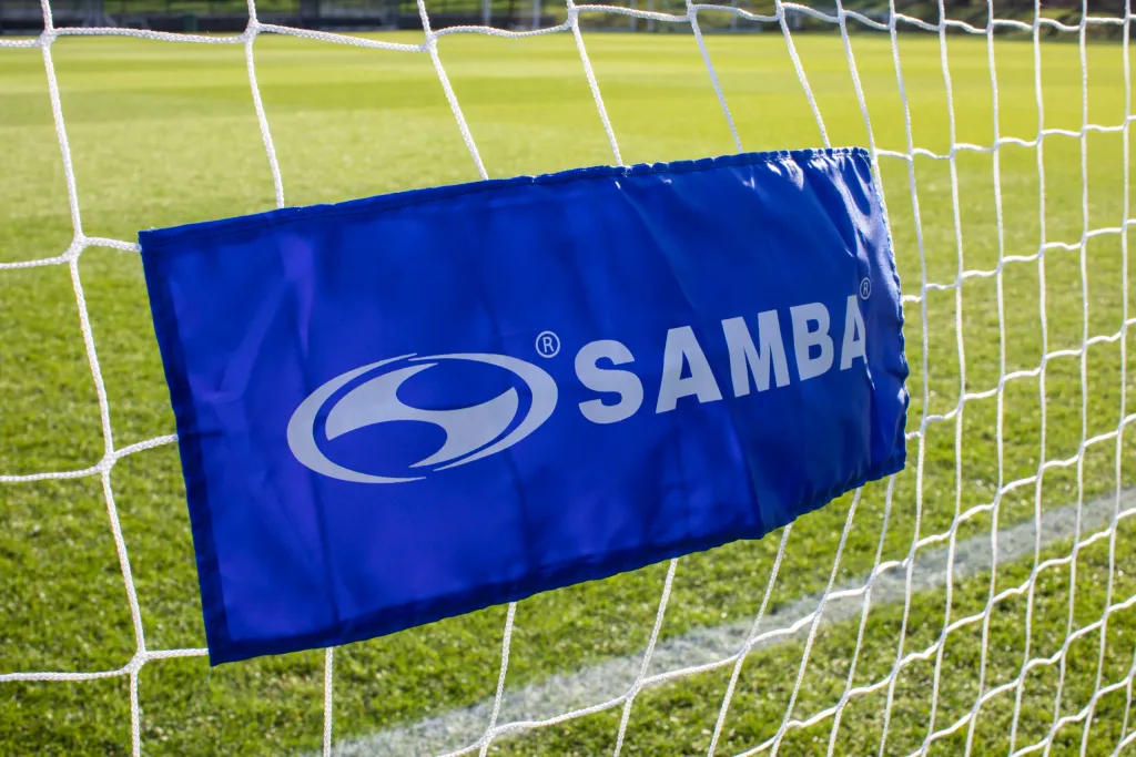 Blue banner reading SAMBA with a white logo attached to a white soccer goal net over a grass field.