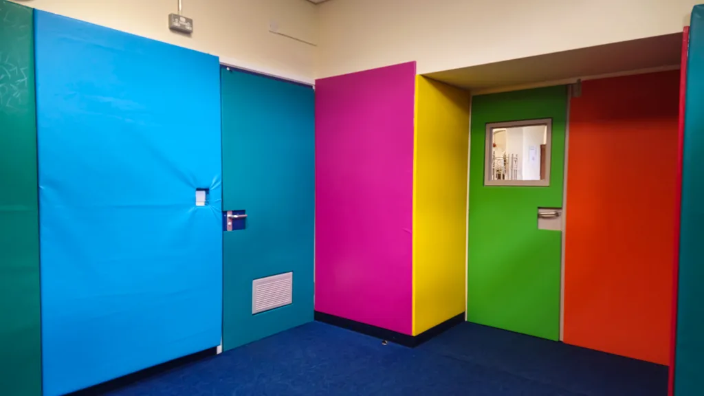 Colorful padded door panels in blue, teal, magenta, yellow, green and orange covering doors and walls, the green door features a small window.