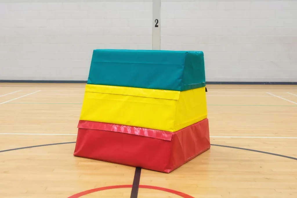 Three-layer vaulting box with red base, yellow middle and green top in PVC-coated nylon with carry handles, placed on a gymnasium floor.