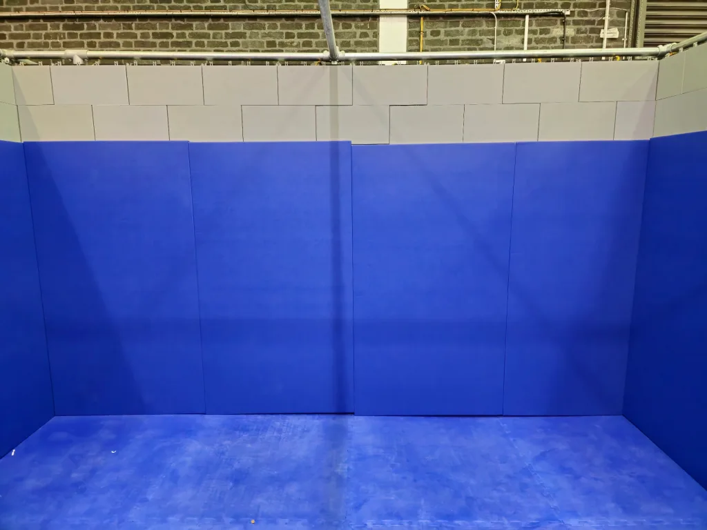 Blue padded floor and walls forming a training corner with light gray foam blocks above and exposed brick and pipes at the top.