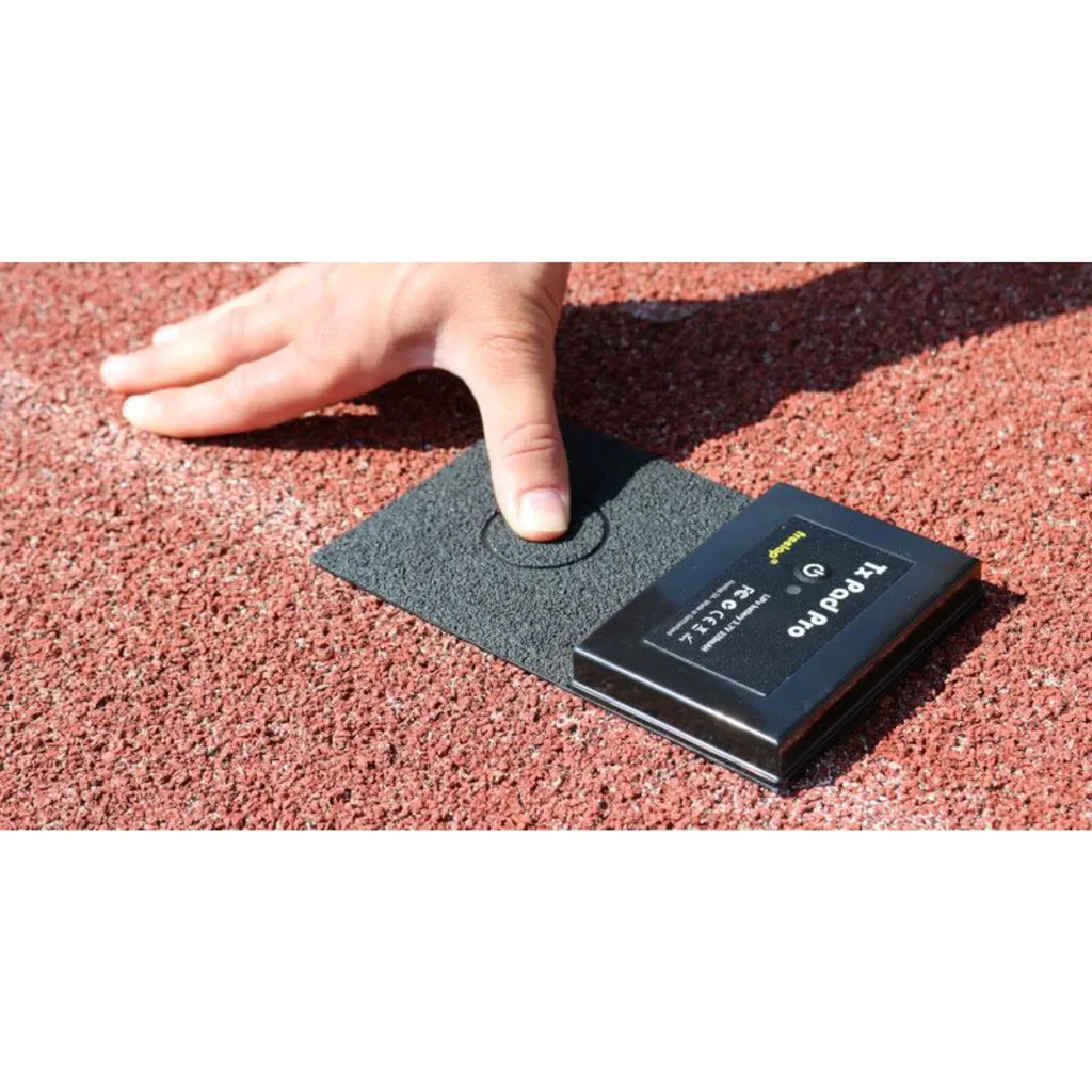 A hand presses a black TX Pad Pro transmitter on a red running track.