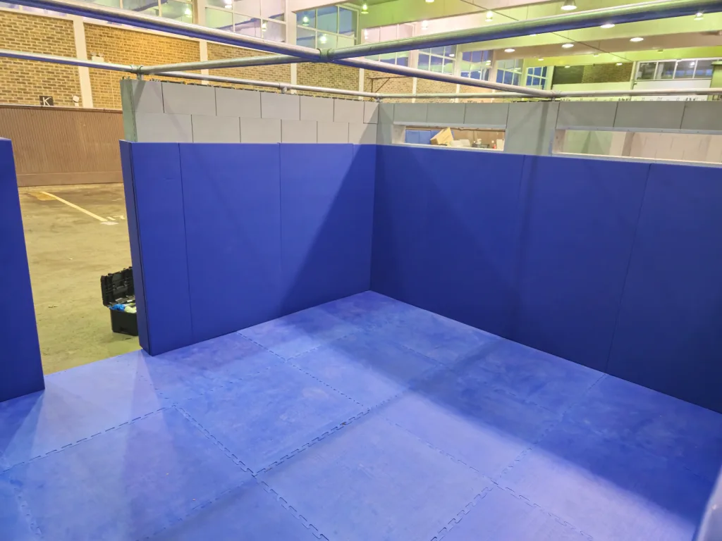 Blue foam wall pads and interlocking floor mats forming a corner training area inside an indoor facility.