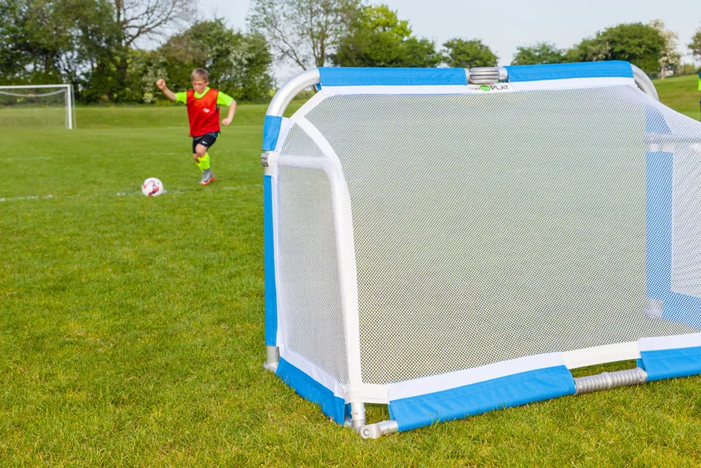 Folding aluminium football goal with white mesh and blue trim on grass, a child in a red bib kicking a ball toward a distant goal behind it.