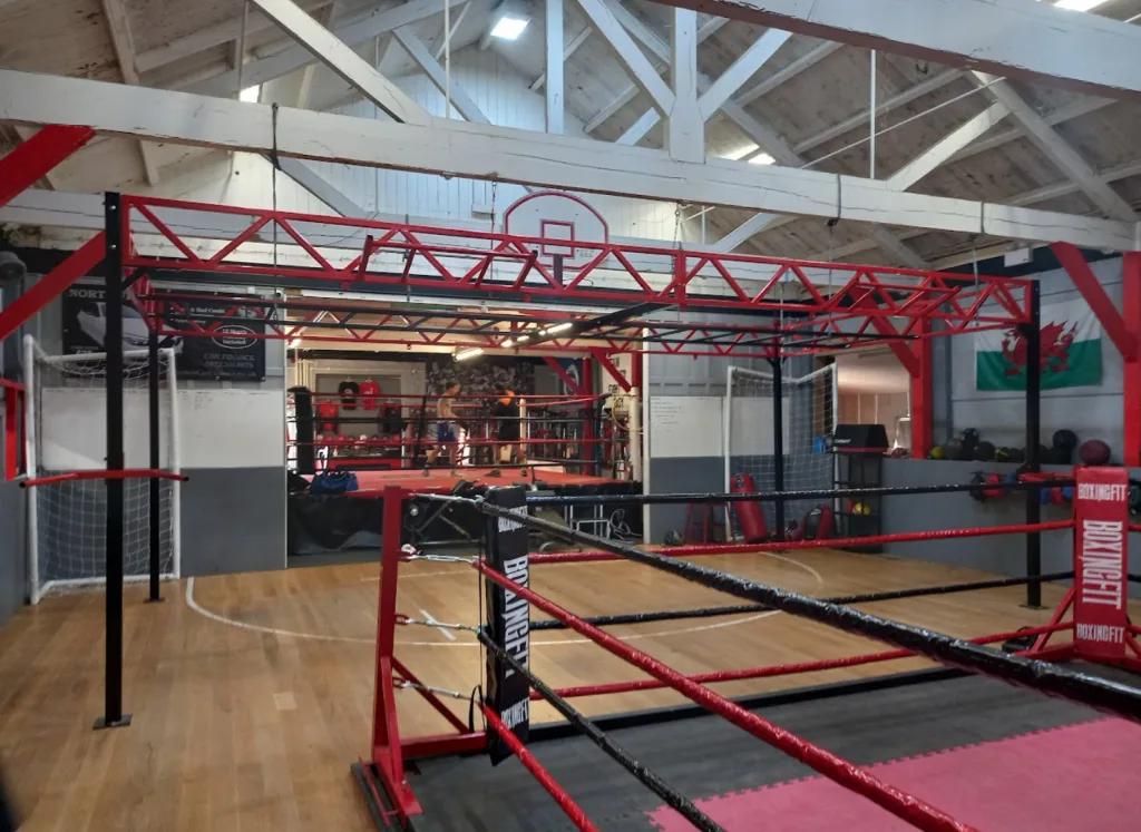 Steel powder-coated equipment rig with overhead truss over a boxing ring in a gym interior, "BOXINGFIT" on the corner pad.