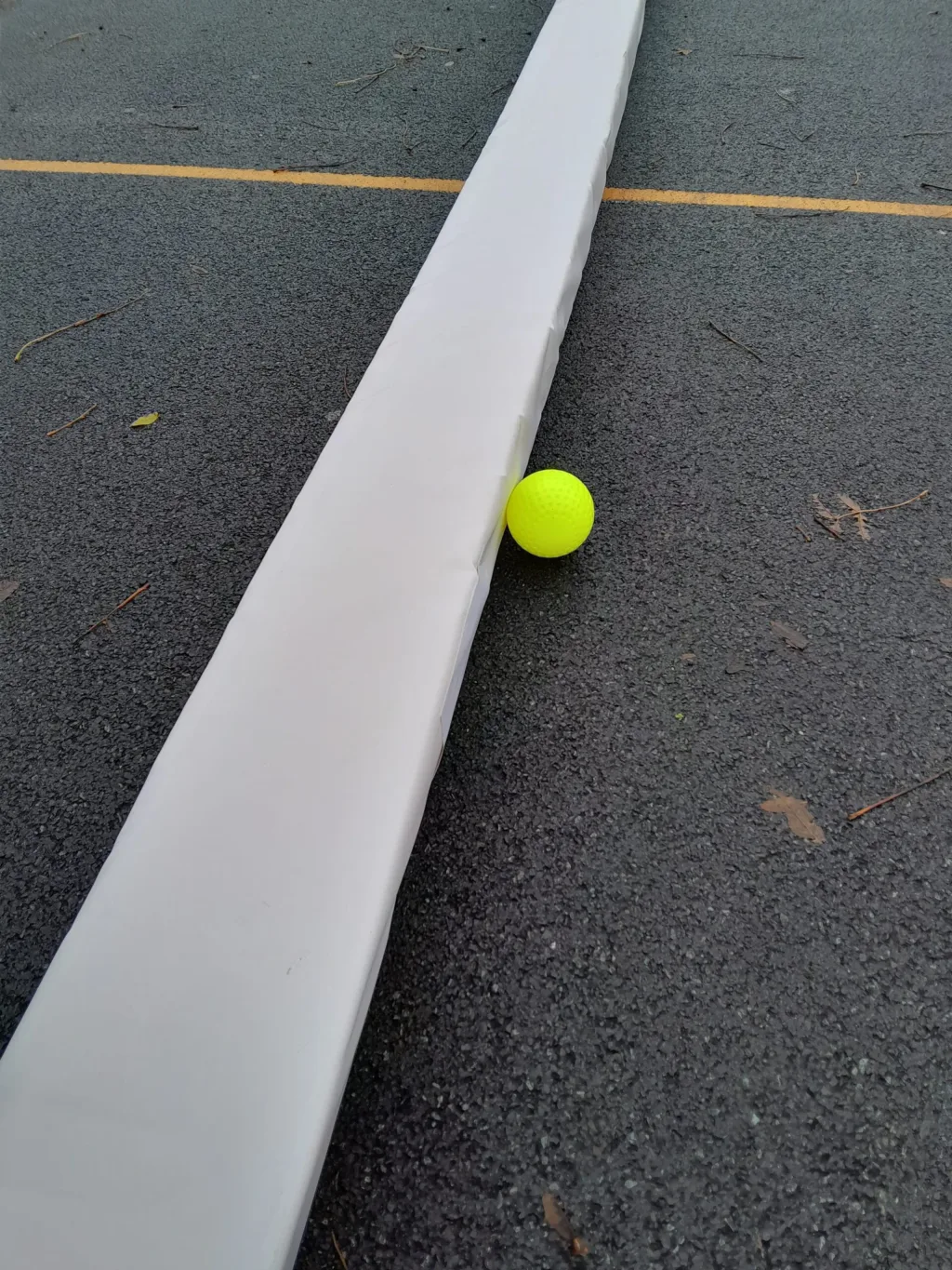Hockey pitch divider pad with sewn heavy duty PVC cover lying on dark asphalt, a bright yellow ball wedged beside it and a yellow court line visible.