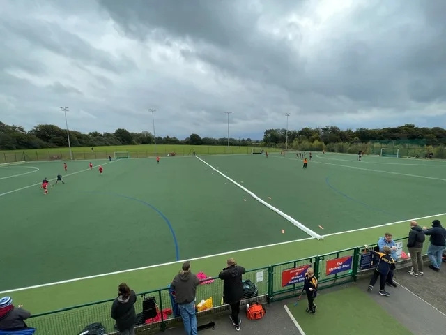 Hockey pitch with green turf split by a long white padded PVC-covered foam divider, players on the field and spectators along a fence.