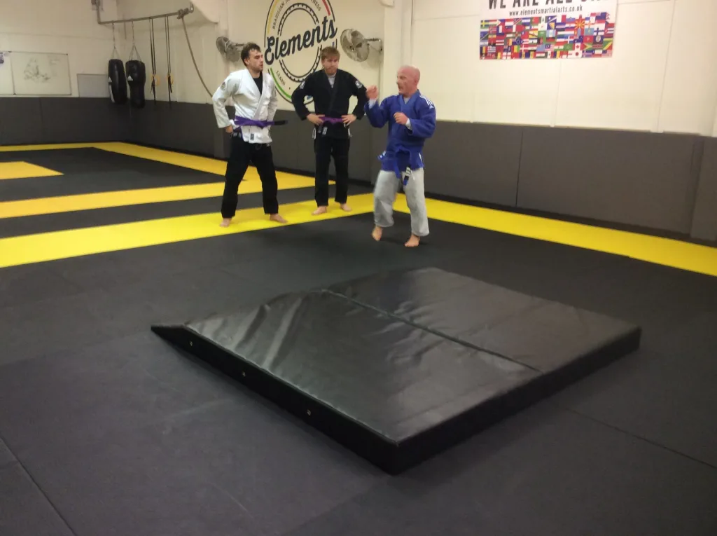 Black PVC-covered slam mat with one end chamfered sits on a dojo floor, three people in gis stand behind and a wall sign reads "Elements".