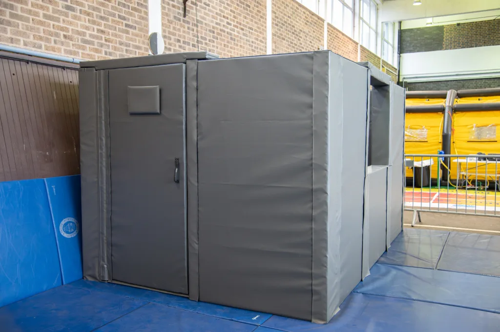 Gray padded foam custody cell module with PVC covering, door with handle and small padded panel, set on blue mats inside a gym hall.