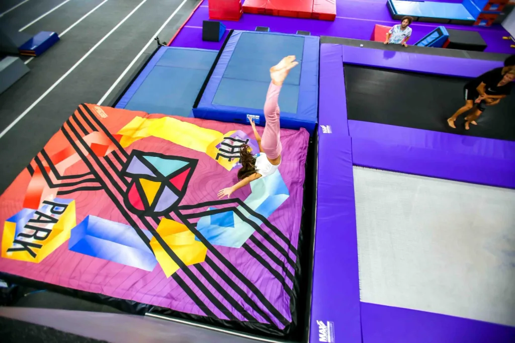 Girl flipping headfirst onto colorful airbag printed with geometric shapes and the word PARK, next to purple trampolines.