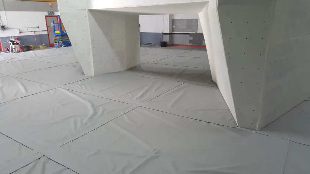 Joined PVC bouldering mats with visible seam lines covering the floor under white climbing wall supports in an indoor gym.