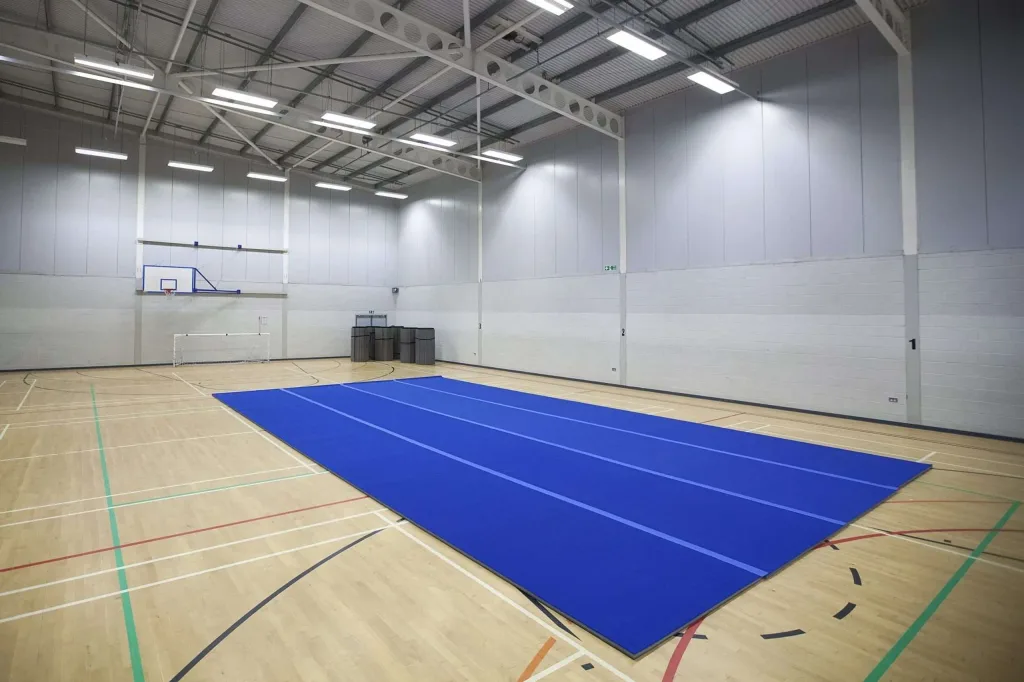 Blue carpet roll-out mat with lighter-blue lane lines laid on a wooden indoor sports hall floor under a raised basketball hoop.